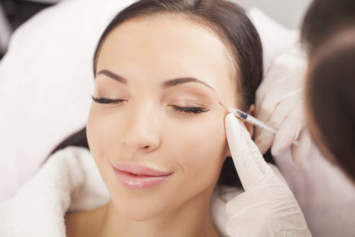 Attractive healthy woman is getting botox injection and smiling. Her eyes are closed with pleasure. The expert beautician is filling hyaluronic acid into female face with syringe